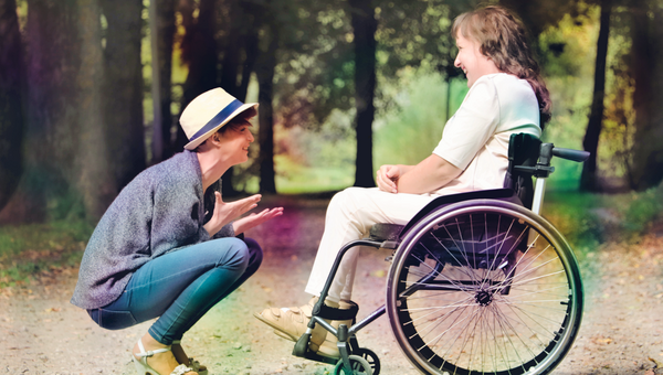 Frau mit Hut in der Hocke spricht mit Frau im Rollstuhl