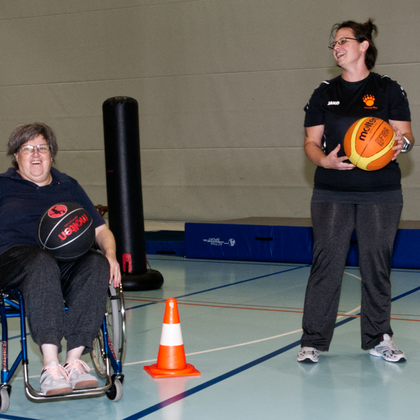 Zwei Personen in einer Turnhalle, beide halten einen Basketball, eine Person sitz in einem Rollstuhl